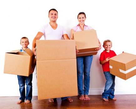A family of four, including two young children, standing indoors against a plain white wall, each holding a large cardboard box. The father and mother are centrally positioned, smiling at the camera, with the father holding a large, empty box in front of him and the mother carrying a smaller box stacked on top of a medium box. The two children, a boy on the left and a girl on the right, each hold smaller cardboard boxes, with the boy's box partially open and the girl’s box sealed with packing tape. The family is dressed casually, with the father in a white t-shirt and the mother in a pink checkered shirt. The scene takes place on a wooden floor, suggesting an interior setting, possibly part of a house move in the Brompton area or nearby postcode in London, and reflects activities related to house removals and packing for a relocation, which Brompton Man with Van services often assist with. Natural lighting illuminates the space, emphasizing the cardboard boxes, with minimal background detail other than the plain white wall.