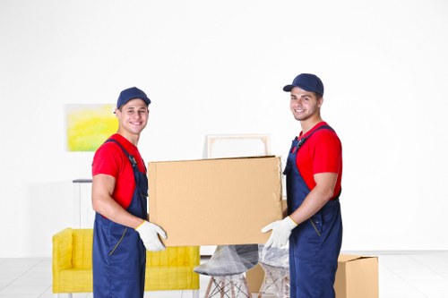 Two removal specialists from Brompton Man with Van, dressed in red shirts, blue overalls, and white gloves, are indoors in a well-lit room with a white background. They are smiling and holding a large cardboard box together, positioned at waist height. Behind them is a yellow sofa with a green and yellow abstract painting mounted on the wall. To the right, a small cardboard box rests on the floor, and a stool draped with a grey cloth is visible. The scene suggests a professional house removal operation, focusing on careful handling of household items, in an environment consistent with typical residential interiors. The clean, minimalist setting emphasizes the meticulous process of packing or preparing items for relocation, highlighting Brompton Man with Van's expertise in sustainable and efficient moving services within the local area near Brompton, London. The overall impression is one of professionalism, care, and routine activities involved in domestic removals.