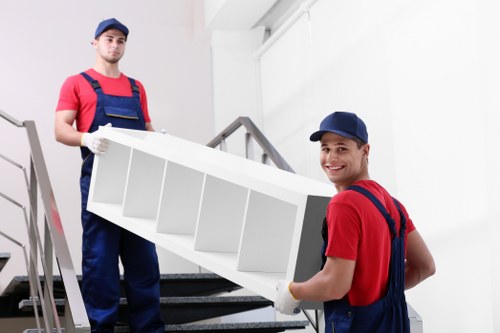 Two professional removal workers, dressed in red t-shirts, navy overalls, and blue caps, are positioned on a residential staircase with a white interior. The worker in the foreground is smiling and looking towards the camera while holding a white, multi-shelved furniture piece with both hands. The second worker is standing behind him, also gripping the furniture securely, and is looking directly at the camera. The staircase has dark steps and a metal bannister on the left side, and the background features a bright, clean, white wall with natural lighting illuminating the scene. This setting is typical of a house or flat interior, and the workers are preparing to transport the furniture item, reflecting a professional house removal or relocation service, possibly around Brompton or the surrounding area in London. The image emphasizes careful handling, teamwork, and efficient moving practices common in Brompton Man with Van’s removals services.