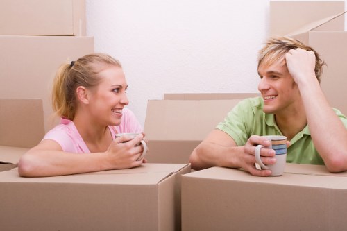 A young woman and a young man with light skin are inside a residential property, sitting among various cardboard boxes used for house removals in Brompton. The woman, with blonde hair tied in a ponytail, is wearing a pink top and holds a ceramic mug, smiling as she looks at the man. The man, with short blonde hair and wearing a light green t-shirt, is also smiling, resting his head on his left hand while holding a mug in his right hand. The background shows a plain white wall and several stacked cardboard moving boxes, some partially open, indicating an ongoing relocation process. The scene is well-lit with natural or artificial light, emphasizing the casual and positive atmosphere typical during a home move. The arrangement of boxes suggests they are preparing to load or unload furniture or personal belongings as part of a professional house removal service in the Brompton area.