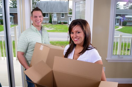 A young man and woman inside a bright, modern house with large windows overlooking a suburban street in Brompton, holding and carrying empty cardboard boxes ready for house removal. The man, dressed in a light green checkered shirt, is smiling while standing behind the woman, who is wearing a white top and also smiling. The background reveals a well-maintained lawn and neighboring houses, emphasizing a residential area in Brompton. The household items, primarily cardboard boxes, are positioned centrally, with the woman holding one box towards the camera, indicating an active moving process. The environment features natural daylight, enhancing the clarity and neutral tone of the scene, which aligns with professional house removal services offered by Brompton Man With Van. The image captures the typical step of packing in a home transition within Brompton, illustrating a friendly, professional approach to relocations in this London suburb.