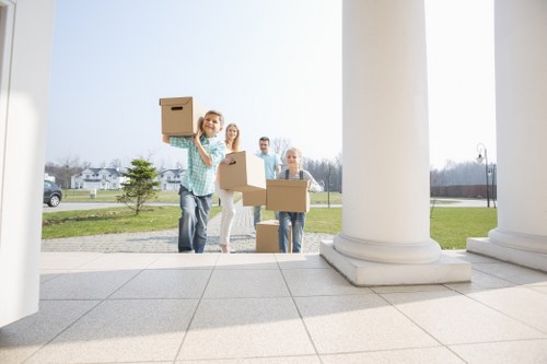 A group of four people are standing outdoors on a paved area of a residential property in Brompton, with a traditional house featuring white columns and a step in the foreground. They are holding cardboard boxes, indicating they are in the process of moving or packing. The individuals include a young woman in a blue plaid shirt, smiling and carrying a box above her shoulder, and three others behind her, holding additional boxes. The background shows a green lawn, a few trees, and a street with parked cars, under a clear sky. The scene captures a moment of teamwork during a house move, with no visible furniture or appliances, only cardboard packing boxes. The location aligns with Brompton’s typical suburban environment, supporting local SEO relevance. Brompton Man with Van, a house removals specialist, appears to be involved in local residential relocation and packing services, demonstrating skilled handling of household items in a typical neighborhood setting. The image emphasizes community-based moving activities in a calm, outdoor suburban context.