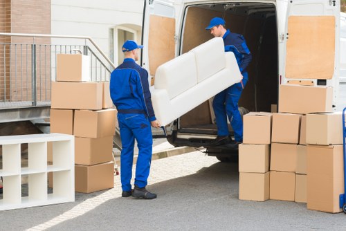 Two professional movers from Brompton Man with Van, dressed in blue uniforms and caps, are engaged in loading a large, cream-colored sofa into a white removals van parked on a residential street. The van's rear doors are open, revealing the inside workspace for furniture loading. One mover, standing inside the van, is carefully lifting and guiding the sofa, while the other stands outside, assisting with the placement. Surrounding the van are several stacked cardboard boxes of varying sizes, some neatly arranged on the pavement, indicating a systematic packing process. To the left, a small white shelving unit with a few empty compartments adds to the scene context. The environment appears bright with natural daylight, and the background features a residential building with a wooden railing and brick exterior. This scene captures the precise moment of furniture loading during house removal services in Brompton, highlighting careful handling of household items and the organized process of a professional move in a suburban setting.