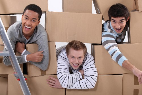 Three young men are inside a room filled with stacked cardboard boxes, which appear to be part of a house removal process. The man on the left, with short dark hair and wearing a grey top, is smiling broadly and leaning on the boxes. In the center, a man with light brown hair, wearing a white and navy striped shirt, is lying on his stomach on top of several boxes, smiling at the camera. To the right, another man with dark hair and a striped long-sleeve shirt, is reaching forward and smiling, partially emerging from behind the boxes. The background shows more stacked cardboard boxes, indicative of a residential interior, possibly a hallway or room during packing or unloading. Bright, natural lighting illuminates the scene, emphasizing the casual, organized environment typical of house removals in a London suburb, such as Brompton or nearby areas. Brompton Man with Van provides professional removals, and this image depicts the friendly, cooperative team involved in household moving activities.