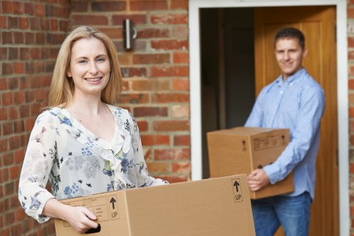 A woman with long blonde hair, wearing a white blouse with blue floral patterns, is standing outside a brick-built house, smiling and holding a medium-sized cardboard box with 'UP' arrow symbols printed on it. Behind her, a man with short dark hair in a light blue shirt is also smiling and carrying another cardboard box, slightly larger, towards the open front door of the residence. The scene captures a moment of house removal or relocation, with the two individuals engaged in moving belongings, standing on a driveway or pathway leading to the entrance. The house features a wooden door frame, and the environment appears to be a residential neighbourhood, consistent with Brompton or nearby areas in London. Visible household items include the cardboard boxes, suggesting packing or unpacking activity typical of local removals services undertaken by Brompton Man with Van. The lighting is natural, indicating a daytime setting, and the overall scene is professional and orderly, emphasizing careful handling of personal belongings during a home move.