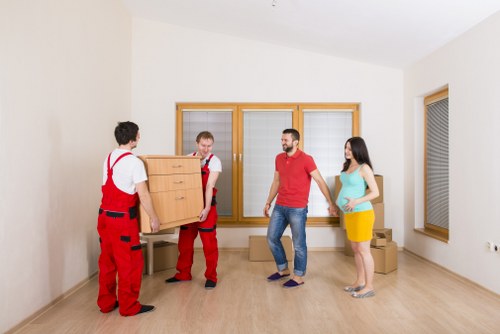 In a bright, minimally furnished room with light wooden flooring and white walls, two movers dressed in red overalls are carefully carrying a wooden dresser with multiple drawers, while a man in a red T-shirt and jeans along with a woman in a blue top and yellow skirt observe. The room features large windows with wooden frames, allowing natural light to fill the space, and a cardboard box is visible on the floor near the woman. The scene likely depicts a house move in the Brompton or nearby area, illustrating the process of relocating household furniture and belongings as part of a professional residential removals service by Brompton Man with Van. The environment is clean, organized, and emphasizes careful handling of furniture during a typical home transfer, aligned with the company's focus on safety and efficiency during house removals in London or southwest London areas.