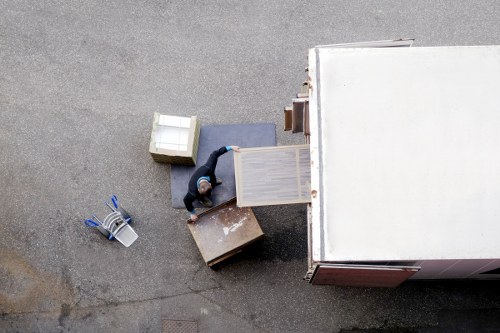 An aerial view of a male removal worker inside a moving van parked on a residential street near Brompton. The worker is lifting a wooden side table onto the van's open rear door, which reveals an interior packed with various household furniture and boxes. In the foreground, a small bedside table with a white drawer, a larger cardboard moving box, and a grey upholstered piece are visible. To the left, a metal basket containing packing supplies such as tape, scissors, and bubble wrap is scattered on the asphalt. The scene captures the process of loading home belongings, with the van positioned adjacent to a house or flat entrance that is off-frame. The street surface appears clean, and natural daylight illuminates the scene, emphasizing the textures of the furniture's wood and fabric, as well as the cardboard boxes and metal tools. This image reflects typical house removal activity in the Brompton area, demonstrating careful handling of household items for a local residential move undertaken by Brompton Man with Van.