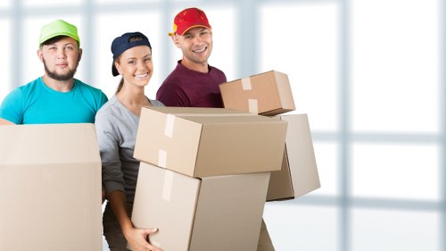 The image shows three professional house removals staff standing indoors in a well-lit area, possibly within a residential property in Brompton or nearby. They are smiling and appear to be in the process of a relocation, each holding or surrounded by cardboard boxes of various sizes, all sealed with packing tape. The boxes have a plain brown finish with visible creases and tape strips, indicating they are ready for transport or packing. The team members are dressed in casual work attire suitable for moving: the man on the left wears a turquoise T-shirt and a green cap, the woman in the middle is in a gray top with a navy baseball cap, and the man on the right wears a maroon shirt with a red cap. The background features large windows with natural daylight streaming in, illuminating the scene and giving a neutral, professional atmosphere typical of a house removal service environment. The setting suggests a residential space in or near London, consistent with Brompton or the SW10 postcode area, emphasizing the company's focus on local house removals services in London.