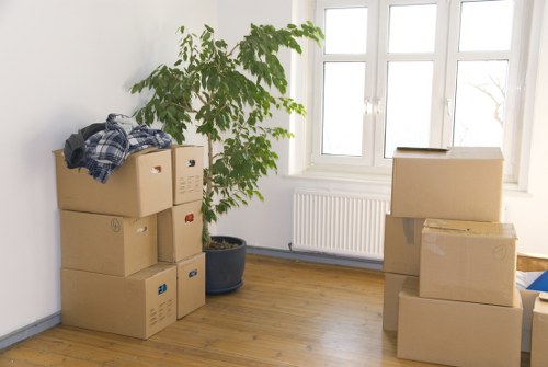 The image depicts a well-lit room with white walls and a wooden floor, featuring several cardboard moving boxes stacked in two separate groups near the walls. The boxes vary in size and are sealed with packing tape, with some labeled or marked. On the left side, there is a small potted plant with lush green leaves positioned next to a few boxes, adding a touch of greenery to the space. Behind the plant, there is a large window with multiple panes allowing natural daylight to illuminate the room, and beneath the window, a white radiator is visible. The room appears to be part of a residential property in a suburban or town setting, possibly within Brompton, as inferred from the postcode and local context. The boxes and minimal furniture suggest a packing or moving process is underway, consistent with house removals services in the area, with a focus on preparing household items for transportation. The overall scene conveys a moment of transition, typical of professional removals in London or surrounding boroughs, emphasizing the organization and careful handling of belongings during a move.