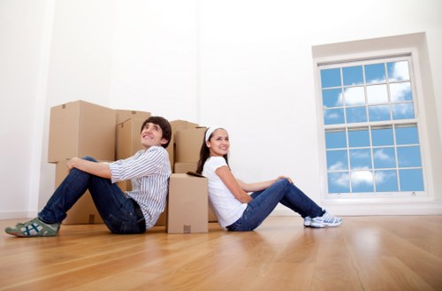 A young man and woman sit on a polished wooden floor inside a bright, empty room near a large double-glazed window with white frames, showing a clear blue sky with some clouds outside. The man, dressed in casual striped shirt and dark jeans, leans back against a stack of medium-sized cardboard boxes with a relaxed expression. The woman, wearing a white top and light blue jeans with white sneakers, sits with her legs extended forward, smiling gently. The cardboard boxes are arranged behind them, indicating they are in the process of moving or packing. The room has white walls and minimal decor, with natural light illuminating the space. This scene is typical of house removals or relocation activities, subtly reflecting a residential setting in the UK, possibly in Brompton or nearby, associated with professional house moving services by Brompton Man with Van.