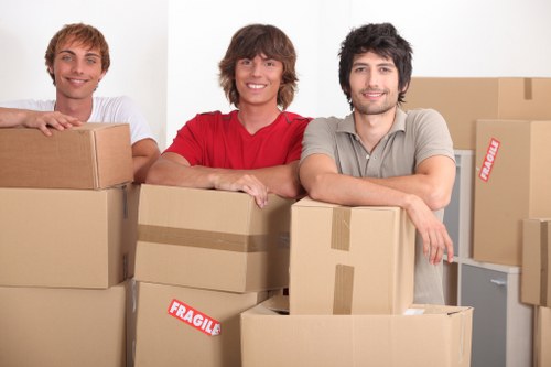 A group of three young men with light skin and casual clothing are standing behind several cardboard boxes in a residential interior, likely a hallway or living room, with some boxes stacked and others at different angles. The man on the left, wearing a white t-shirt, has short, curly hair and is smiling. The man in the middle, with medium-length brown hair, is wearing a red t-shirt and also smiling. The man on the right, with dark wavy hair and a light grey polo shirt, is smiling and leaning on a stack of boxes. The boxes vary in size, some sealed with packing tape, with one visibly marked 'FRAGILE' in red and white labels, indicating they are packed for moving or house relocation. The background shows a plain white wall and a small glimpse of a kitchen or hallway area, with the scene suggesting a professional house removals process, potentially in Brompton or nearby areas. The lighting is bright and even, highlighting the household items prepared for transfer, consistent with the services of Brompton Man with Van in house removals.