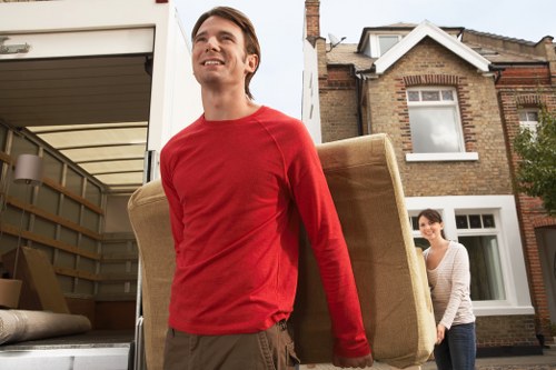 A young man with short brown hair, wearing a red long-sleeved shirt and brown trousers, is smiling and carrying a large, beige upholstered armchair with visible fabric texture, out of a vehicle parked outside a residential house. Behind him, a young woman with dark hair tied back, wearing a light grey cardigan and white top, is assisting by holding the armchair and guiding it towards the house entrance. The house is a typical brick terraced property with white window frames and a small front garden. The vehicle is a white van with an open side door, revealing additional household items and moving blankets inside. The scene takes place in daylight, with natural light illuminating the driveway and highlighting the activity of household relocation, fitting within the context of local house removal services in the Brompton area.