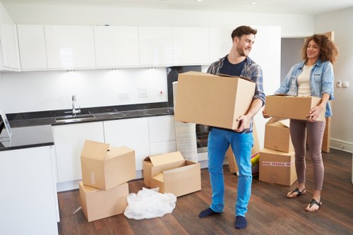 A young man and woman are inside a modern kitchen, engaging in a house removal activity with Brompton Man with Van. The man, wearing a plaid shirt and blue trousers, is carrying a medium-sized cardboard box, holding it with both hands, and smiling as he moves towards the camera. The woman, dressed in a light denim jacket over a white top and maroon trousers, stands near a pile of unpacked boxes on the dark wooden floor, holding an open cardboard box filled with household items. Several cardboard boxes of various sizes are scattered throughout the kitchen, some sealed with packing tape and others open, revealing contents like tissue paper and household items. The kitchen background features white cabinets, a black countertop, and a stainless steel sink, with a light-filled window illuminating the scene, suggesting daytime. The scene captures a typical house relocation process within a residential property in the Brompton area, emphasizing the furniture and packing materials used in local removals services.