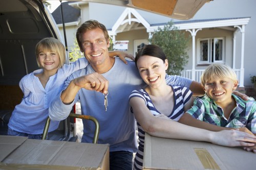 A smiling family of four, consisting of a man, a woman, and two children, engaged in a home moving process outdoors in front of a residential house in Brompton. The man, with short light brown hair and casual attire, is holding a set of keys in his right hand and standing behind an open van trunk, which contains cardboard boxes and packing materials. The woman, with short black hair and wearing a navy and white striped shirt, is leaning on a cardboard box with her left arm. The two children, a girl and a boy, are on either side of the adults; the girl has blonde hair and is dressed in a light blue blouse, while the boy has short blonde hair and is wearing a colourful plaid shirt. The background features the exterior of a white, traditional-style house with bay windows, a small porch, and greenery in the garden. The scene is well-lit by natural daylight, capturing a moment of moving and new beginnings, exemplifying professional house removal services in a residential neighbourhood near Brompton.