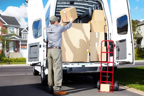 A male house remover wearing a grey shirt, beige trousers, and a cap is loading a small cardboard box into the back of a white moving van parked on a residential street. The van's rear doors are open, revealing several other large cardboard boxes stacked inside, some with visible tape and labels. A red two-wheeled hand trolley with a small cardboard box on its base is positioned nearby on the pavement. The background features modern suburban houses with well-maintained gardens, trees, and a partly cloudy sky, suggesting a typical residential area in Brompton or nearby postcode areas. The scene is well-lit with natural daylight, emphasizing the organized packing activity associated with house removals and local moving services provided by Brompton Man with Van.
