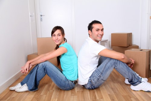 An image showing a young woman and a young man sitting on the light wooden floor of a residential interior, back-to-back with their knees bent and arms resting on their legs. The woman, on the left, wears a blue T-shirt and blue jeans, with long brown hair and white sneakers. The man, on the right, wears a white T-shirt and blue jeans, with dark hair and white sneakers. Behind them are several cardboard moving boxes of varying sizes, some stacked, in a room with white walls and a closed white door visible in the background. The scene depicts a moment during a house move, with the boxes prepared for transportation, and the overall environment appears clean and well-lit, emphasizing the context of residential removals in the Brompton or nearby area.