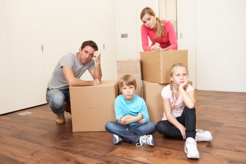 A family of four, comprising a man, a woman, a boy, and a girl, appears to be in a residential interior, preparing for a move as part of house removals services. The man, dressed in a light grey T-shirt and jeans, is crouched next to a large cardboard box, resting his head on his hand. The woman, wearing a pink top, is leaning over several stacked cardboard boxes, possibly packing or organizing belongings. The children are seated on the wooden floor; the boy, wearing a blue T-shirt and jeans, looks directly at the camera with a neutral expression, while the girl, in a white T-shirt and dark pants, rests her chin on her hand, appearing contemplative. The room has plain white walls and minimal furniture, suggesting an empty or decluttered space in a house in Brompton or nearby. The hardwood flooring adds warmth to the setting, and the scene is illuminated by natural or ambient light, emphasizing the household items and the family’s activity related to relocation preparations, typical of Brompton Man with Van’s removal services.