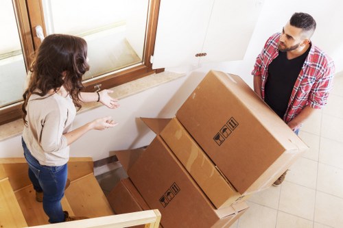 A man and a woman are indoors during a house move, standing on a tiled entryway floor near a staircase with wooden steps. The woman, with long dark hair, is dressed in a light-colored top and dark jeans, and is gesturing with her hands towards three large cardboard boxes stacked on the staircase. The boxes are sealed with packing tape and have handling symbols printed on the sides. The man, with short dark hair and a beard, is wearing a red and white checkered shirt over a black t-shirt, and is holding the top box from the side, preparing to carry it. The environment appears well-lit with natural light coming from a nearby window, and is part of a residential property in Brompton. Brompton Man with Van's removal service is implied through the scene, illustrating a typical home relocation process.