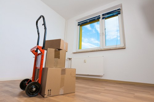 The image shows a residential room with light-colored wooden flooring and plain white walls, featuring a large double window with blue and yellow striped blinds, partially open to reveal a view of a bright, blue sky with some clouds. In the corner of the room, a stack of three cardboard moving boxes, varying in size, is positioned near the wall beneath the window. The boxes are sealed with packing tape, with the largest box at the bottom and the smallest on top. To the left of the boxes, a red and black hand truck (dollies) with two wheels and a handle is leaning against the wall, ready for use. The room appears clean, with minimal furnishings, suggesting it is at the beginning or end of a house removal process. The scene subtly indicates local moving activity, possibly in Brompton or nearby areas, consistent with Brompton Man with Van's services in the removals sector, within a typical home environment in the UK.