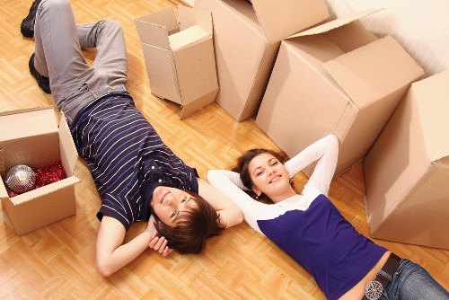 Two young women are lying on a wooden floor inside a residential property, surrounded by open cardboard boxes indicative of a house move. The woman on the left has short brown hair and is dressed in a dark navy and white striped shirt with grey trousers; she is lying on her back with her hands resting behind her head. The woman on the right has long brown hair and is wearing a white top with a blue sleeveless dress layered over, also lying on her back with arms behind her head, smiling gently. The boxes around them vary in size, some filled with packing materials such as bubble wrap and shredded paper, and are positioned along the walls, suggesting a recent or ongoing unpacking process. The room appears well-lit with natural light, and the overall scene conveys a moment of rest during a house relocation, consistent with the services provided by Brompton Man with Van in the Brompton area.