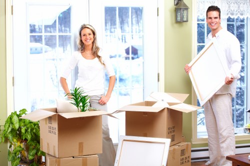 A smiling woman with long, wavy blonde hair, wearing a white long-sleeved top and light grey trousers, stands indoors near large windows with white frames, behind several open cardboard moving boxes of varying sizes, some filled with household items and a small potted green plant with long, narrow leaves. To her right, a man with short dark hair, dressed in a white shirt and light trousers, holds a picture frame or flat item, also smiling. The scene is set in a bright, well-lit room with a light green wall on the right side, and the windows reveal a wintery outdoor scene with snow and houses. Multiple empty packing boxes are stacked around, and a household setting with casual decorations creates a typical home environment appropriate for house removals services in Brompton or nearby areas. The image captures a moment of packing or preparation for a move, reflecting professional house removal activities.