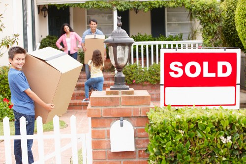 A family of four, including two children, are engaged in packing and moving household items outside a residential property in Brompton, demonstrating house removals services. The young boy in the foreground is holding a large cardboard box, ready for transport, while the girl next to him appears to be supervising or preparing to carry another item. In the background, an adult woman and man are standing near the porch, with the woman holding a smaller box and the man guiding a larger cardboard box as they prepare for the move. The scene is set in front of a house with a white picket fence, brick pillars, a brick pathway, and a black lamppost, with lush green shrubbery and a front garden providing a tidy residential atmosphere. The house features a porch with a white railing and windows, indicating a typical suburban property in the Brompton area. A 'SOLD' sign is prominently displayed in the garden, indicating the property has recently changed ownership, and the overall scene captures a daylight moment of a household transition typical of local removals services in London's southwest region.