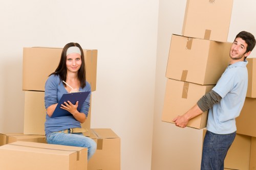 The image depicts a young woman with long dark hair, wearing a blue top and light denim jeans, sitting on top of several stacked cardboard boxes inside a plain room with cream-colored walls, holding a clipboard and pen. To her right, a young man with short dark hair, dressed in a light blue shirt with rolled-up sleeves and dark blue jeans, is smiling while carrying a medium-sized cardboard box with visible tape securing its edges. Several other cardboard boxes of various sizes are arranged randomly around them, some stacked and others placed on the floor, indicating they are in the process of packing or moving. The setting appears to be a residential interior, possibly a living room or hallway, with no furniture or decorations visible beyond the boxes. The natural lighting suggests daylight, and the scene reflects a typical house removal or relocation scenario, as might be offered by Brompton Man With Van services in the Brompton area or nearby TW postcode region, aimed at facilitating local household moves safely and efficiently.