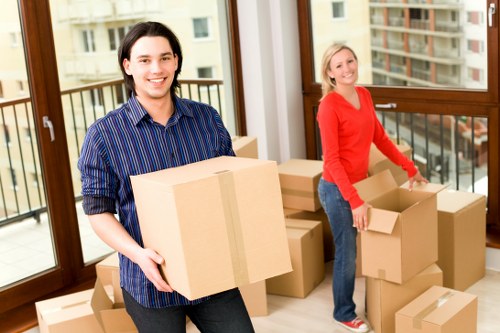 The image depicts two individuals inside a modern residential property with large floor-to-ceiling windows and a balcony, preparing for a house move. The person in the foreground is a young man with dark hair, wearing a blue striped shirt, smiling at the camera while holding a cardboard box sealed with packing tape. Behind him, a woman with blonde hair in a red long-sleeved top and jeans is also smiling as she works with partially filled cardboard boxes, some open and some taped shut, arranged on the wooden floor. The boxes contain household items, though specific contents are not visible. The environment features natural daylight illuminating the space, highlighting the clean and bright interior with a neutral colour palette. Outside the windows, part of an urban street scene with buildings is visible, suggesting a town setting. The scene captures a typical interior of a house or flat in Brompton, with a focus on the process of packing and organising belongings for a removal, fitting within the context of professional house removals services offered by Brompton Man with Van in the area.
